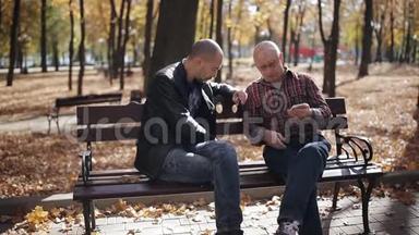 一位年迈父亲的成年儿子在秋公园的长凳上教智能手机。